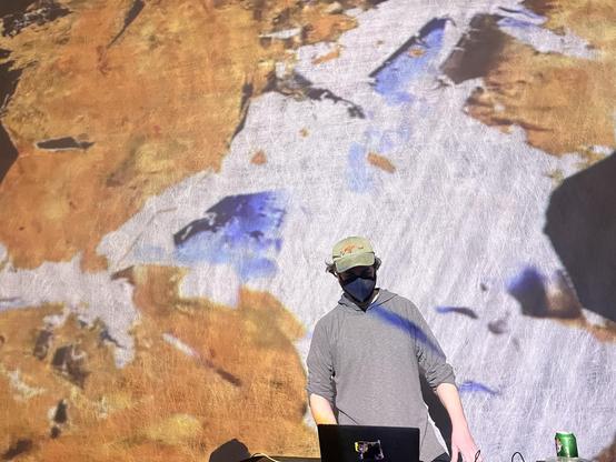 performance pic from Algorithmic Art Assembly in 2022 - a man in a green hat with a coati, grey sweater, and black covid mask performs on a laptop in front of a large projection screen with brown, white and blue glitchy video collage