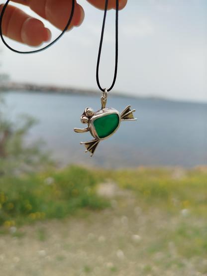 a silver frog pendant with a green semitransparent gemstone. it's hanged from black cord