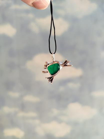 a silver frog pendant with a green semitransparent gemstone. it's hanged from black cord