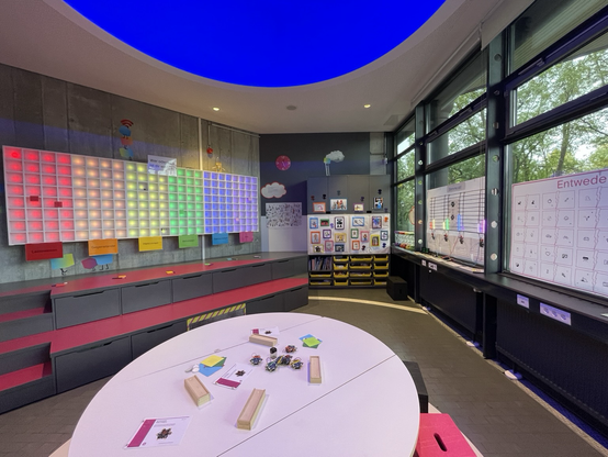 An der linken Wand ist eine Art Setzkasten, der in Regenbogenfarben beleuchtet wird. An Erbe des Raumes ist eine Galerie mit Roboter Fotos. In Vordergrund ist ein Tisch, auf dem Bastelmaterial liegt.