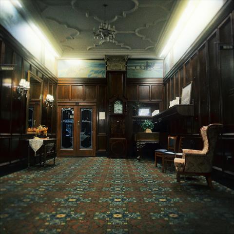 A large hallway resembles a mansion's study, complete with wall sconces, a wingback chair, a grandfather clock, and double-doors leading to a ballroom in the distance.
