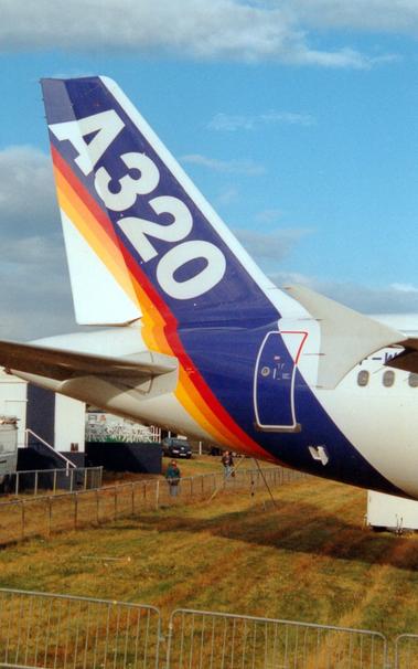 Close-up of the tail and rear fuselage of a twin engined jet airliner parked facing to the right.
The plane is largely white, with a thick dark blue diagonal band around the tail and rear fuselage, followed by thinner red, orange, and yellow bands.
There are large, white, "A320" titles on the blue band.
Part of the registration "F-W" is visible between the wing and the grey winglet on the right of the frame.
Grass fills the bottom of the frame, with a temporary metal fence surrounding it, and a couple of people looking up at the plane, with temporary buildings behind them.
Blue sky with fluffs of white fill the rest of the frame.