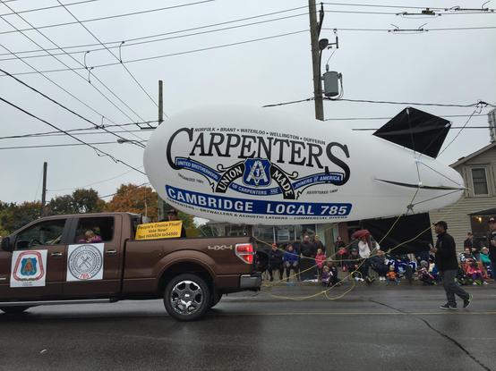 A brown pickup truck on a wet urban street. It has a dirigible-shaped balloon floating about its bed. The balloon reads, in part:

“Norfolk, Brant, Waterloo, Wellington, Dufferin Carpenters

United Brotherhood of Carpenters and Joiners of America

Cambridge Local 785”