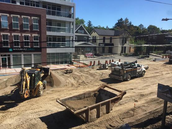 A bird’s-eye view of street construction in an urban setting. The street is all dirt/earth. There’s a front-end loader moving earth from a large pile. There’s a hydro truck. In the background there’s a mid-rise apartment building and a houses. The sky is clear and blue.