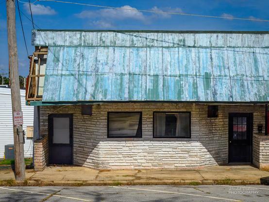 The front of a one-story stone-faced office building.  There are two recessed entrance doors, two windows, and a blue/green faded metal roof with a piece missing at the left end of the building.
