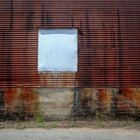 A portion of the side of a very rusty metal Quonset Hut building with a window opening that has been covered by a piece of white sheet metal.