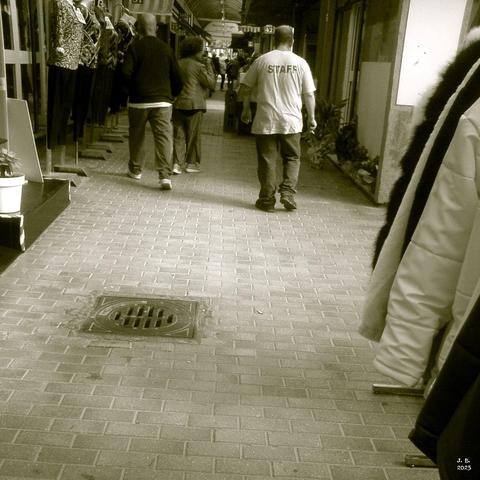 A monochrome sepia photo of people walking away in an indoor market in Rome, Italy. One sturdy man wears a white t-shirt with the word STAFF written on the back.