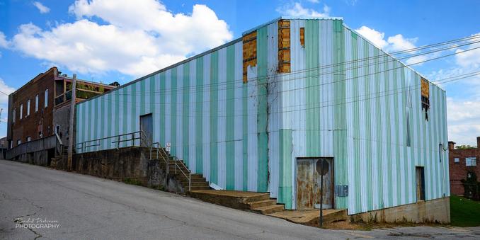 An old brick building is covered with stripes of green and white metal siding.  A concrete wall/sidewalk/stairs connect a door on the left side of the building to street level and another door at the mitered corner of the building.
