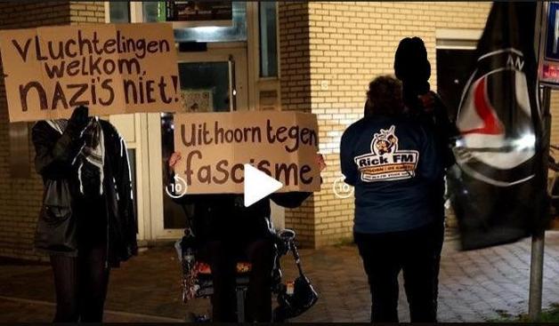 Drie antifascisten by night in Uithoorn. Twee staande personen, 1 met kartonnen bord voor het gezicht en 1 met Afa vlag, waarvoor een verslaggever vanaf de rug gezien staat, (met een logo op de rug) en in het midden een persoon in rolstoel met kratonnen bord voor het gezicht. De teksten: vluchtelingen welkom, nazis niet. Uithoorn tegen fascisme