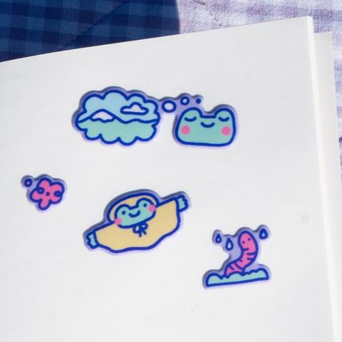 Photograph of a stickers in a stickerbook. The stickers are puffy so a bit 3D and show a variety of small rain themed icons: A frog dreaming of the mountains, a red flower, a worm looking up from the grass and a frog in a raincoat.