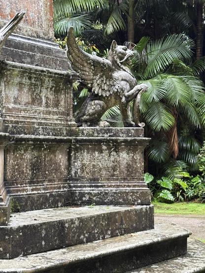Statue of a lion with a moustache and wings sitting on its haunches.  Forms one of four stone animals guarding a larger monument.