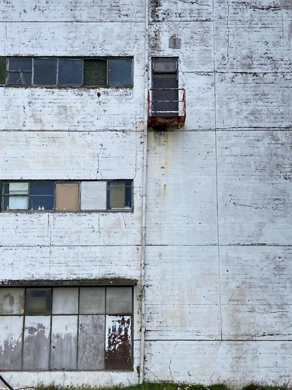 A very small rusty balcony on a run down building with broken windows and cracked concrete.