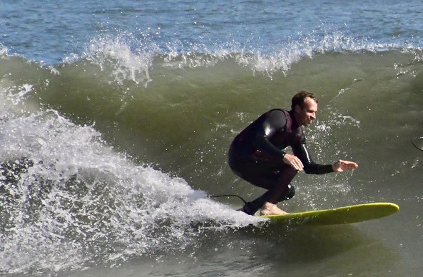 Surfing smiling while catching a wave on a yellow surf board. Surfer is wearing a black wetsuit. A green wave is curling behind him.