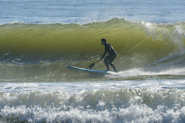 a person paddle surfing — standing on a blue paddleboard while riding a large, greenish wave. The surfer is wearing a full black wetsuit and holding a paddle, maintaining balance as the wave curls dramatically behind him