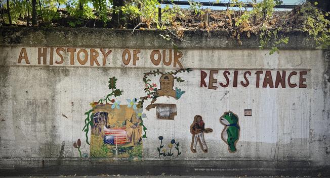 A weathered concrete wall transforms into a powerful canvas of community storytelling, where vibrant painted murals declare "A HISTORY OF OUR RESISTANCE" in bold rust-colored letters. The artwork celebrates resilience through diverse imagery: Indigenous figures adorned with traditional headdresses intertwine with colorful vines and flowers, creating a living tapestry of cultural preservation. A vintage American flag fragment emerges from layered textures, while small human figures stand as testament to collective strength.

The striking emerald frog sitting prominently on the right side of the mural carries special significance - its bright green skin and freshly painted appearance directly references the viral Portland ICE protester Seth Todd, who sparked a nationwide movement by donning an inflatable frog costume to peacefully resist immigration enforcement policies. This 24-year-old activist chose the frog specifically to counter narratives of violent extremism, stating "the best way to demonstrate that is by wearing a frog costume" as he danced outside Portland's ICE facility. The mural's frog serves as a symbol of creative, nonviolent resistance - embodying the spirit of protesters who used whimsical animal costumes to challenge the Trump administration's characterization of Portland as a "war zone".​

Above the mural, wild vegetation cascades over the wall's edge - dried brown vines and fresh green foliage create a natural crown, while glimpses of vehicles peek through 