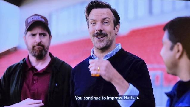 Brendan Hunt, Jason Sudeikis, and Nick Mohammed watching some training out on the pitch in the pilot episode. Jason is smiling holding a clear glass of Gatorade. He tells Nick, "You continue to impress."