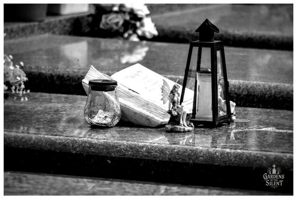 A black and white close up photo shows several commemorative objects resting on a polished granite surface, likely a tombstone or memorial.

From left to right: a small, empty, clear glass jar with a black lid; a stack of paper or perhaps a small, open book; a small figurine of a winged fairy seated near the center; and a tall, slender black metal lantern with glass panes.

The objects are in sharp focus, while the background, which contains flowers, is softly blurred.