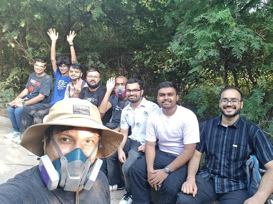 A person wearing a camel-colored travel hat and a blue reusable respirator, taking a selfie with 8 men sitting behind per, some of them gesturing happily at the viewer.
