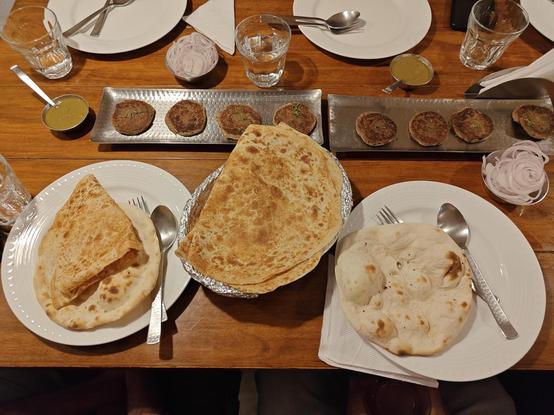 Photo of a wooden table. It has two steel trays containing 4 galawati kababs each, served with two cups of smooth mint chutney and two cups of sliced onions. There are also four white ceramic plates, one containing an ultā tawā parānthā and another containing a rumālī rotī. There's a basket between them containing more rumālī rotīs.
