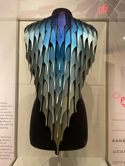 back armor shaped like a cape of fluted, multicolored metal strips that resemble scales, on a slender black mannequin torso.