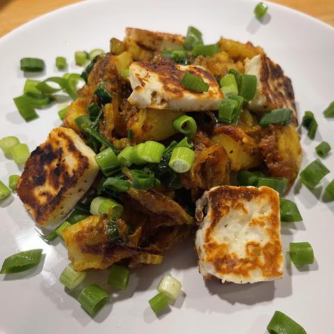 Na białym talerzu porcja ziemniaków w curry z selerem naciowym, szpinakiem i kawałkami grillowanego sera halloumi, posypana świeżą cebulką dymką.