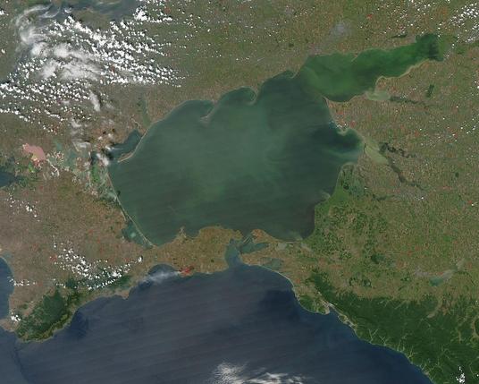 Satelitenaufnahme des Asowschen Meeres, das von grünen Uferlandschaften und landwirtschaftlichen Flächen umgeben ist. Die Wasseroberfläche erscheint in verschiedenen Grüntönen und das Bild zeigt auch Wolken über dem Meer sowie einige helle Punkte, die möglicherweise landwirtschaftliche Flächen oder Siedlungen darstellen.