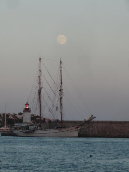 Im Hafen von Ibiza liegt ein Zweimaster am Ende eines geschäftigen Tages. Direkt über einem der Maste prangt der Vollmond am abendlichen Himmel.
