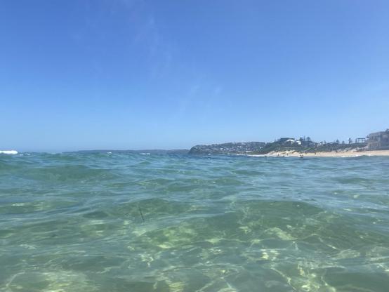 Looking southwards while swimming not far from shore at Bar Beach. Water dominates the view, a clear pale turquoise green, full of refracted light. Light sand, green vegetation and buildings are visible further along the beach. Blue sky and early afternoon light.