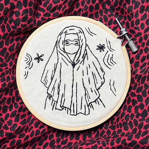 Una obra bordada presenta una figura con hiyab y mascarilla, rodeada de líneas onduladas y pequeños elementos decorativos. El bordado se presenta en un bastidor circular sobre un vibrante fondo de leopardo rojo y negro.