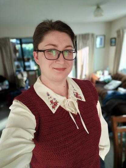 A light-skinned person with short medium-brown smiling into the camera. They are wearing glasses, a super cute cream blouse with embroidered red and purple floral motifs on the collar, and a hand-crocheted sweater vest in a lucious berry red. The background is blurred and unexciting, but shows the photo to have been taken indoors.
