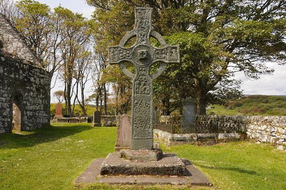 The Kildalton Cross. The image shows part of a ruined church on the left of the frame and a graveyard occupying much of the lower part of the frame with trees behind it. There are only a few scattered graves while, in the centre foreground and occupying the whole central strip of the frame is a large greenish-grey decorated stone cross with arcs connecting its arms to the upper and lower parts. The scene is in sunlight.