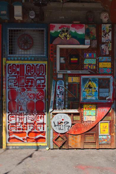 This is a vertical image of a vividly decorated, weathered wooden wall, at the Metelkova autonomous cultural center in Ljubljana. The surface is a chaotic collage of paintings, graffiti, stencils, and found objects. On the left is a door covered in bold, red and white graphic designs, resembling a mix of ornate lettering and abstract shapes. To the right, a wooden surface is layered with small, colourful paintings and frames of various sizes. Notable elements include a skateboard mounted vertically, a circular white sign with red 'Hip Hop' text, and a grinning, carved white face near the top right. The entire scene is bathed in bright, directional sunlight that creates deep shadows, emphasising the rough textures and the intense, saturated colours of the street art.