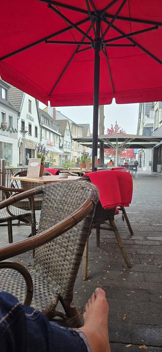 Blich in die Rheinbacher Hauptstraße vom Bäcker Lennartz aus. Oben rote Sonnenschirme unten leere Tische und Sessel. Auf einigen Sesseln liegen rote Decken. Im Vordergrund der nackte Fuß des Fotografen, der in einem der Sessel sitzt und das Bild aus Tischchen aufgenommen hat.