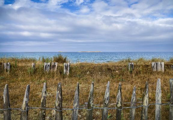 Blick über eher ruppige Vegetation auf das Meer. Im Vordergrund die Spitzen eines sehr einfachen Holzzaunes aus einzelnen spitzen Pflöcken. Er sichert ein kurz bewachsenes Schutzgebiet aus trockenen Gräsern und ein paar kleinen Sträuchern. Vor einer Erhebung stehen ein paar verwitterte Holzbohlen aufrecht, meist in 2er bis 3er Gruppen. 
Dahinter dann das sich kräuselnde Meer. Am Horizont ist eine Insel erkennbar. Der Himmel ist bewölkt mit ein paar blauen Flecken am oberen Bildrand.