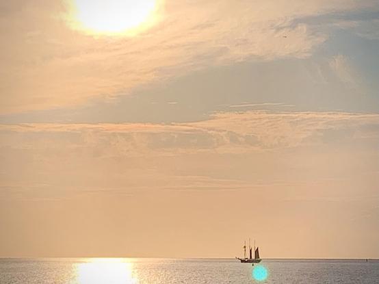 A tranquil seascape at sunset featuring a sailboat in the distance, silhouetted against a softly lit sky with shades of orange and blue. The sun reflects off the water's surface, creating a serene atmosphere.