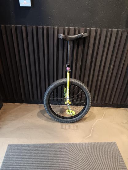 A green framed unicycle leaning against a black wall with a concrete floor.