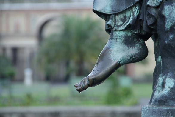 Regentropfen am Fuß einer Bronzestatue. Im Hintergrund das rote Mauerwerk der Orangerie und der grüne Garten.