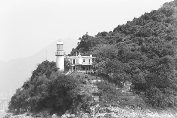 This is a black and white photo in landscape format of the Green Island Lighthouse, Hong Kong (2015).
Stretching from the lower left corner diagonally upwards to the upper right corner is steep rising terrain covered in lush, dense and dark tropical vegetation. Running horizontally along the base of the image is a rocky shore with numerous large pale rocks and just a hint of water visible in the lower left corner. To the left of centre is a medium height (17m), bright white lighthouse, located above a small cliff. It has a cylindrical tower with a large round lantern made of many rectangular panes of glass and finished with a domed top. Beside and to the right is a white, two story house with a flat roof and a verandah surrounding the building. Further right and down the sloping terrain is a garden area with a few small outbuildings. In the far distance and in the upper left diagonal of the photo is a very hazy view of the rising terrain of the New Territories and the top of one of the 300m towers of the Stone Cutters Bridge. Above is an equally hazy sky, possibly clear or possibly overcast.