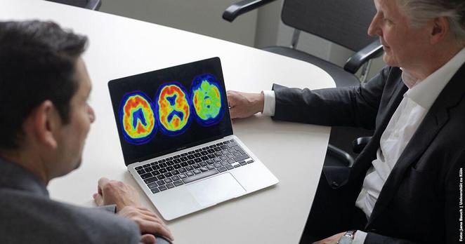 Laptop showing colorful brain scan images on screen, discussed by two seated individuals in formal attire at a meeting table.