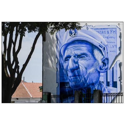 a large mural painted on a wall, depicting the face of an older man wearing a sailor's cap. The mural is rendered in shades of blue, giving it a striking and monochromatic appearance. The text on the mural includes the words "Especialidade" and "Bacalhau," which is Portuguese for "specialty" and "codfish". The mural is set against a backdrop of a white wall, with a tree on the left framing the image. (draft by mistral.ai, revised by author)