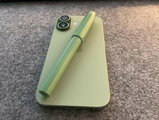 Ein Smartphone in Grün liegt auf einer strukturierten Oberfläche. Das Telefon ist rechteckig mit abgerundeten Ecken. Auf der Rückseite befinden sich zwei schwarze runde Kameras und ein schwarzer Blitz. Ein langer, dünner grüner Stift liegt diagonal über dem Telefon. Der Stift hat an beiden Enden eine schwarze Spitze. Auf dem Stift steht in schwarzer Schrift "Stylus". Die Oberfläche ist grau und strukturiert.