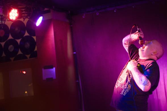 Am rechten Bildrand ein glatzköpfiger Sänger, der nach schräg oben in ein Mikrofon schreit. Die Scheinwerfer, die ihn in violettes Licht tauchen, sind in der linken oberen Bildecke zu sehen.