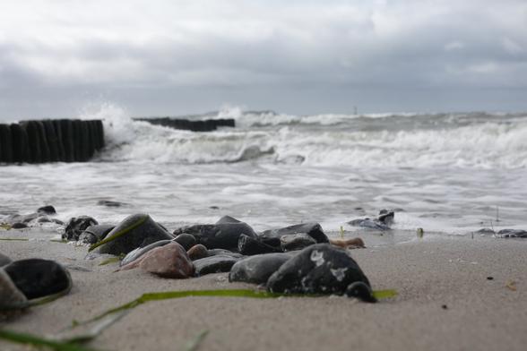 Mehr Steine zu sehen, im Hintergrund bricht sich eine Welle an den Holzbohlen