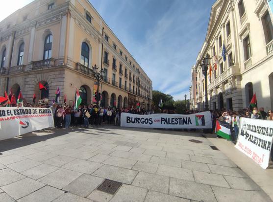 Multitudes de personas con banderas palestinas y vestidas con kufiyas y también camisetas con eslóganes pro palestinos, tras tres pancartas que rodean un espacio vacío – es la Plaza del Cid en Burgos. La pancarta que se ve de frente es la de “Burgos con Palestina” descrita en toot anterior, a la izquierda, contra el estado de Israel y la impunidad de los genocidas; a la derecha “SOLIDARIDAD PALESTINA – Israel estado asesino – con un dibujo de una persona con la cabeza totalmente cubierta con una kufiya y en posición de disparar una honda
