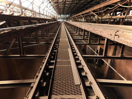 Blick in die Halle der Sinteranlage. Ein langer Gang führt zum Ende der Halle. In der Mitte ein metallener Gehweg, drumherum Gleise. Rechts und links ist die Metallkonstruktion zu sehen. Oben ist das Metallgerüst des Daches.