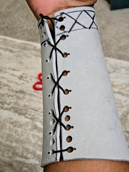A gray EVA foam bracer on a forearm. Black elastic lacing is looped through holes to secure the left and right ends together.