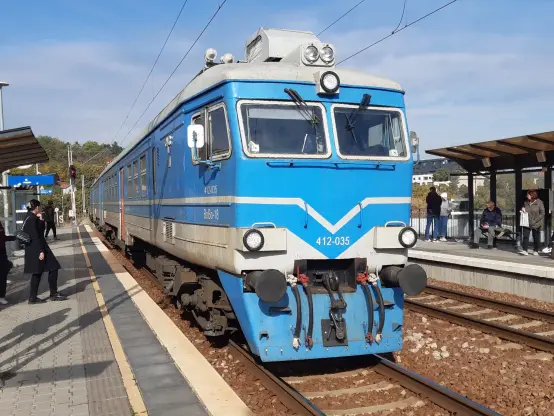 The image shows a blue and white train at a railway station. The front of the train features a prominent driver's cabin with multiple windows and lights. There are passengers on the platform, some standing and others seated, along with benches. The station's surroundings include a clear sky and greenery in the background.