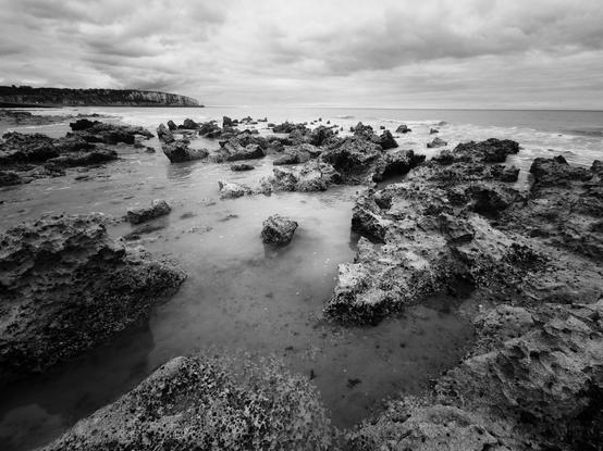 A black and white photograph depicts a rocky coastline with a calm sea. The foreground is dominated by a dense arrangement of dark, irregularly shaped rocks, partially submerged in shallow water. The rocks vary in size and texture, appearing rough and porous. Beyond the rocks, the water extends to a horizon line where it meets a cloudy sky. A landmass, likely a cliff or coastline, is faintly visible in the distance on the right side of the image.