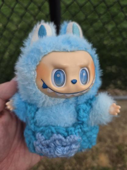 Photograph of a white person's hand holding a blue off-brand Labubu toy, also referred to as a Lafufu, wearing blue overalls made of yarn. Labubu or Lafufu toys have a hard plastic face and hands with a soft, furry body and bunny style ears. The faces have large eyes, a button nose, and fangs from the top of the mouth overhanging their lips in a long smile.