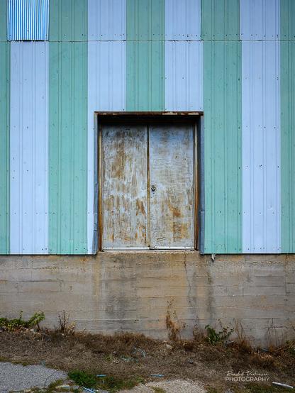 A weathered metal double door on a building wall covered by light green and white striped metal siding.  The door and building are above a tall concrete foundation and there are no stairs to get some to or from the ground about 5 feet below the door.